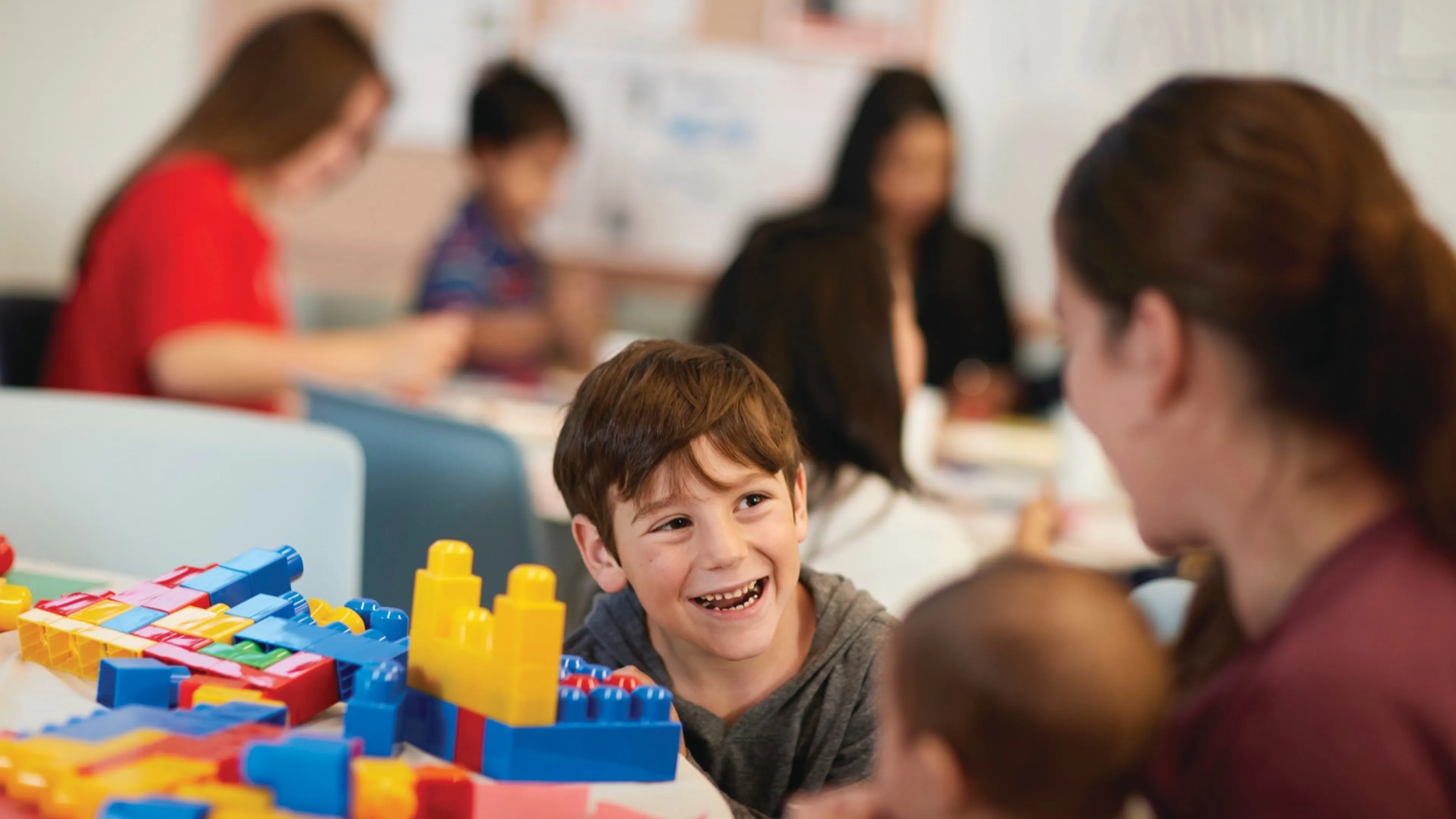 A child smiles while building with colorful blocks alongside an adult.